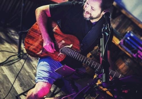 A man is sitting and playing a guitar in a dimly lit setting. There's musical equipment around, and he appears to be performing.