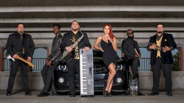 A group of six people posing with musical instruments in front of a car in a parking area.