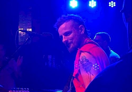 A musician with a red sleeveless jacket and tattoos playing guitar under blue stage lights, facing sideways in an animated performance.