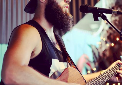 A bearded man wearing sunglasses and a cap plays an acoustic guitar while singing into a microphone.