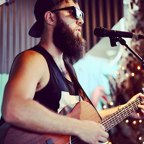 A bearded man wearing sunglasses and a cap plays an acoustic guitar while singing into a microphone.