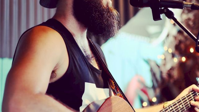 A bearded man wearing sunglasses and a cap plays an acoustic guitar while singing into a microphone.