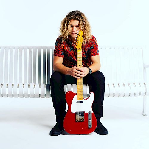 A man with long curly hair sits on a white bench, holding a red electric guitar. The background is plain white.