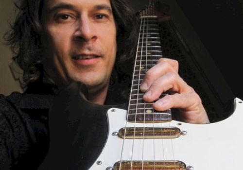 A man with long hair is holding a guitar and smiling while posing for the camera. The guitar is large in the foreground with visible strings.