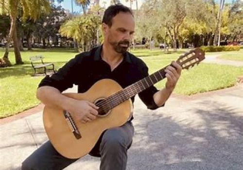 A man is sitting outdoors in a park, playing an acoustic guitar. The background features trees, grass, and a bench.