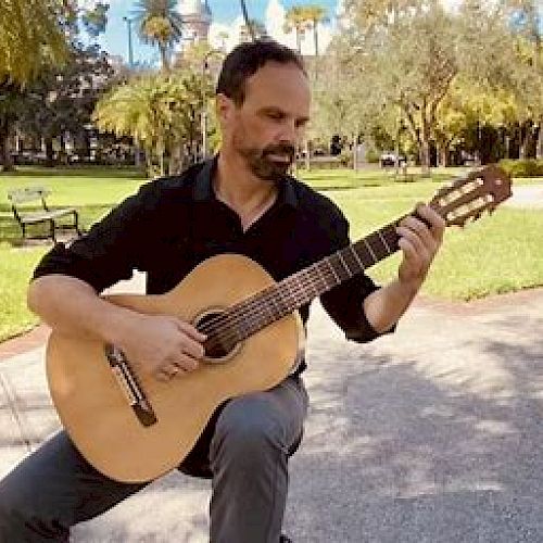 A man is sitting outdoors in a park, playing an acoustic guitar. The background features trees, grass, and a bench.