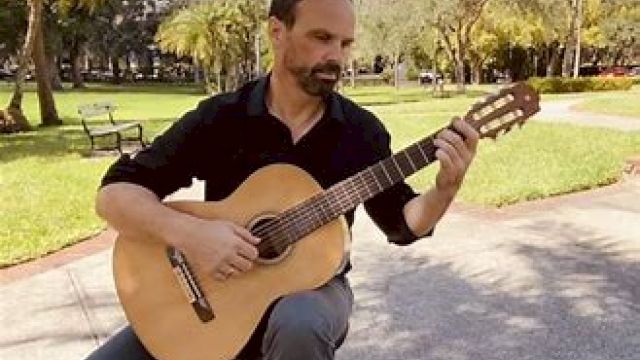 A man is sitting outdoors in a park, playing an acoustic guitar. The background features trees, grass, and a bench.