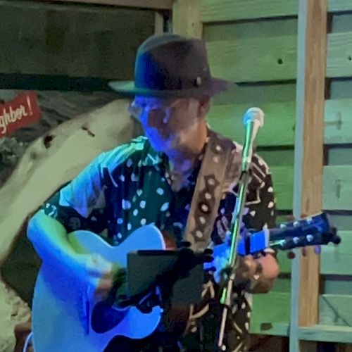 A person wearing a hat, playing an acoustic guitar and singing into a microphone, with a colorful backdrop and wooden structures around.