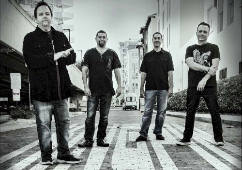 Four men standing on a patterned street in an urban area, with buildings and a vehicle in the background.