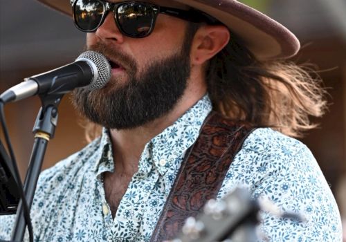 A man with a beard, sunglasses, and a wide-brimmed hat plays a guitar and sings into a microphone.