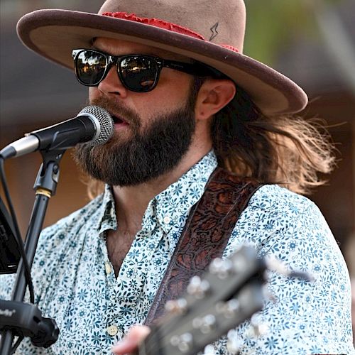 A man with a beard, sunglasses, and a wide-brimmed hat plays a guitar and sings into a microphone.