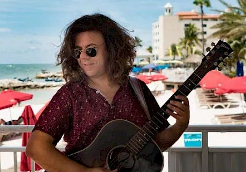 A man wearing sunglasses and a patterned shirt plays an acoustic guitar on a beachfront deck with red umbrellas and a blue sky in the background.