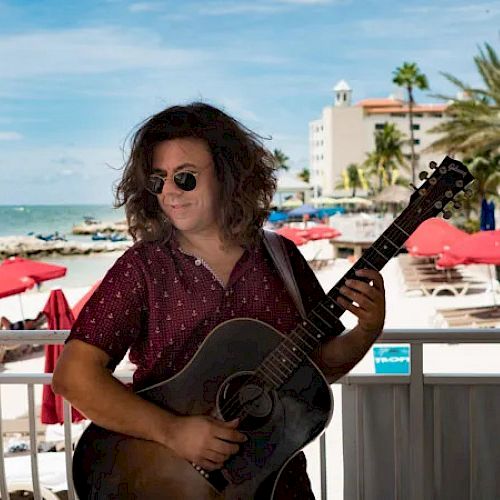 A man wearing sunglasses and a patterned shirt plays an acoustic guitar on a beachfront deck with red umbrellas and a blue sky in the background.