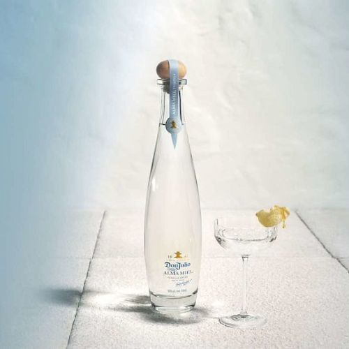 A tall bottle with a yellow label next to a glass containing a lemon wedge on a light-colored surface, creating a minimalist composition.