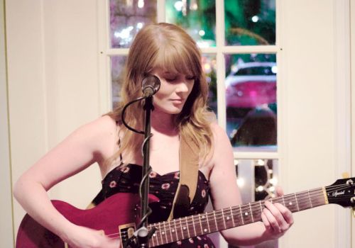 A person with long hair plays an electric guitar and sings into a microphone, standing indoors in front of a window with colorful lights outside the window.