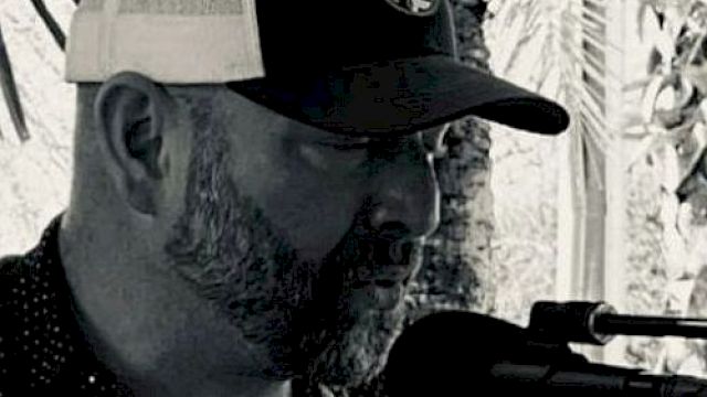 A man wearing a cap and standing in front of a microphone, possibly singing or speaking. The photo is in black and white.