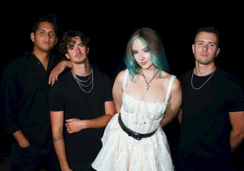 The image shows four people posing together; three are wearing black, and one is in a white dress with blue-green hair, against a black background.
