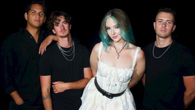 The image shows four people posing together; three are wearing black, and one is in a white dress with blue-green hair, against a black background.