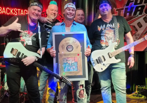 A group of five people with guitars stand on a stage, one holding a plaque, all smiling. There's a drum kit and a banner reading 