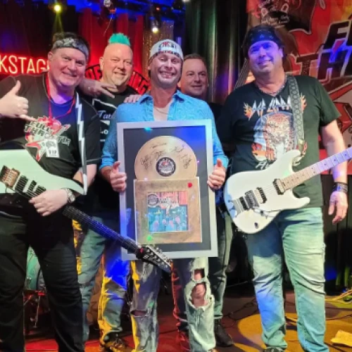 A group of five people with guitars stand on a stage, one holding a plaque, all smiling. There's a drum kit and a banner reading "Full Throttle" in the background.