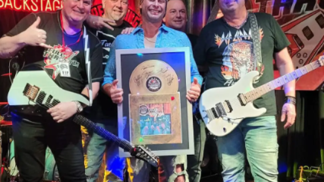 A group of five people with guitars stand on a stage, one holding a plaque, all smiling. There's a drum kit and a banner reading "Full Throttle" in the background.