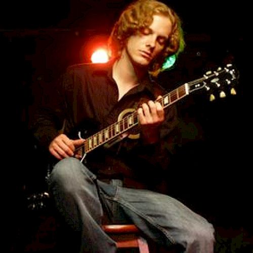 A person with curly hair is seated on a stool, playing an electric guitar under stage lighting.