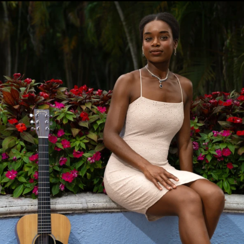 A woman in a light-colored dress sits on a ledge with red and green plants behind her; a guitar rests against the ledge on her left side.