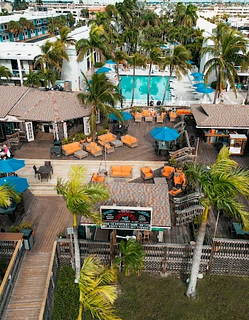 An aerial view of a tropical resort features a pool, palm trees, and outdoor seating areas with umbrellas.