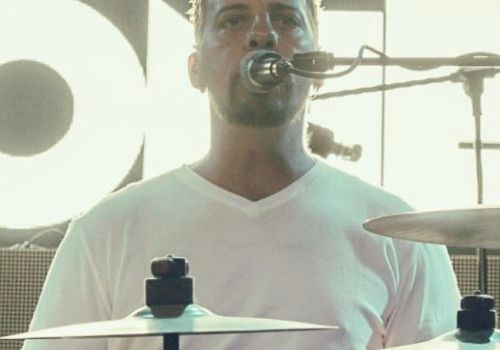 A person is playing drums on stage, wearing a white shirt and singing into a microphone. The background has bright lighting.