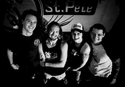 A black and white image shows four smiling individuals standing closely together, each with visible tattoos, under a sign that says 