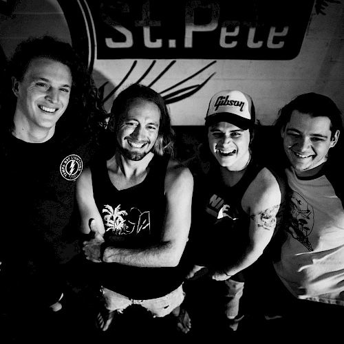 A black and white image shows four smiling individuals standing closely together, each with visible tattoos, under a sign that says 