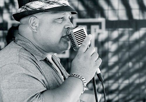 A man wearing a hat sings into an old-fashioned microphone, with a patterned shadow on the wall behind him.