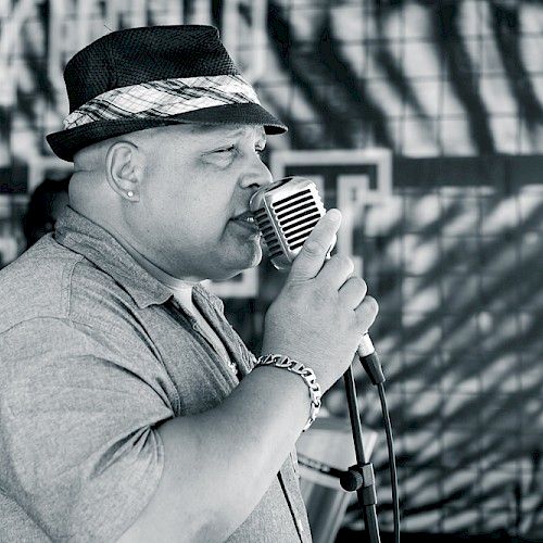 A man wearing a hat sings into an old-fashioned microphone, with a patterned shadow on the wall behind him.