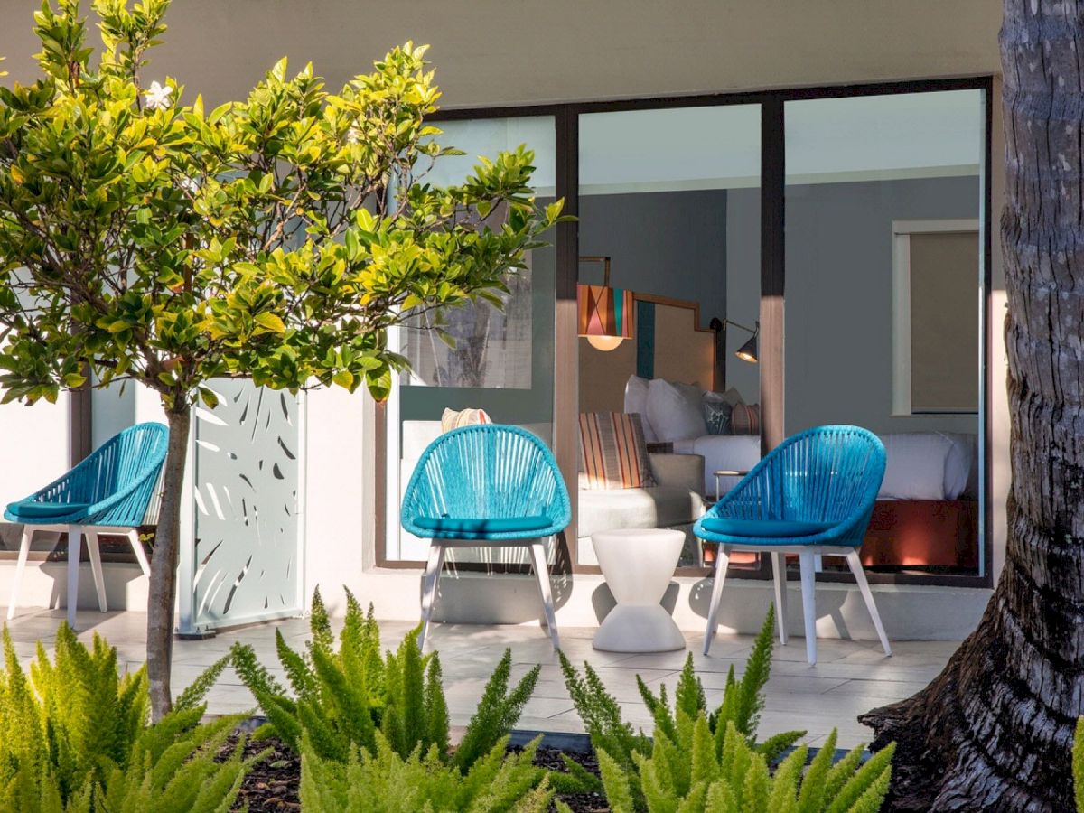 This image shows an outdoor seating area with two blue chairs, a small table, and a view into a room through glass doors, surrounded by greenery.