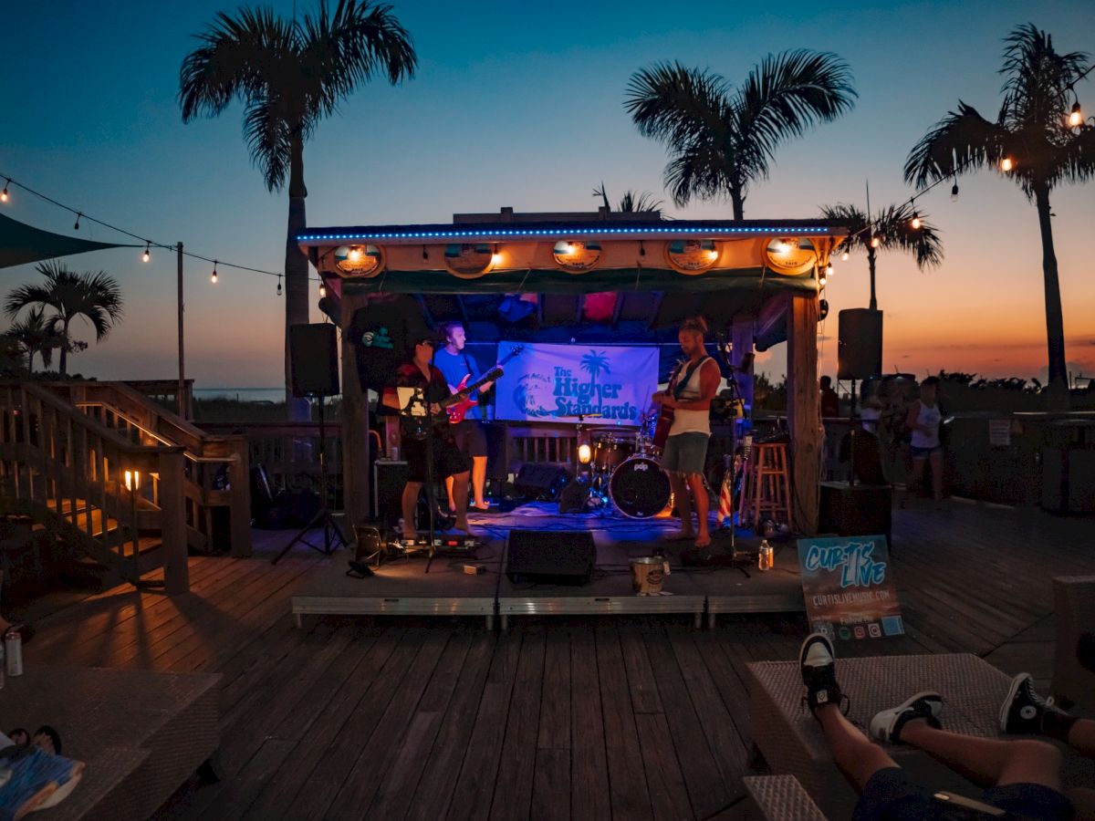 A live band plays on an outdoor stage at sunset, with palm trees and string lights creating a relaxed atmosphere on the wooden deck.