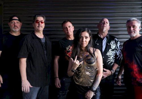 A group of six people posing in front of a dark, ribbed backdrop, with five men and one woman in the center making a hand gesture.