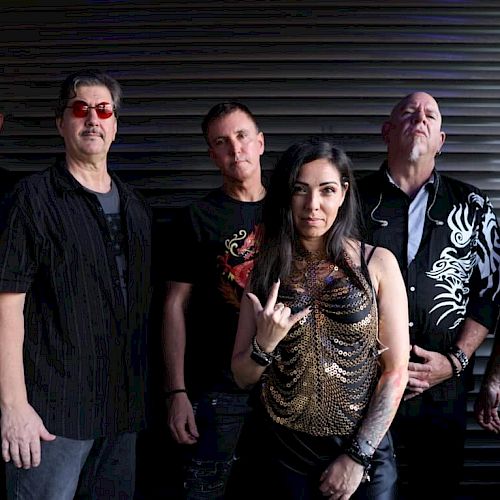 A group of six people posing in front of a dark, ribbed backdrop, with five men and one woman in the center making a hand gesture.