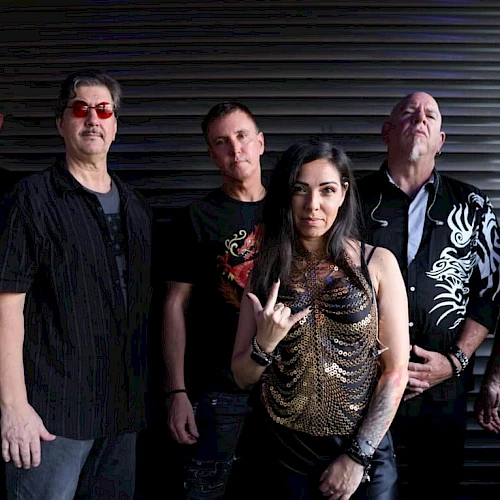 A group of six people posing in front of a dark, ribbed backdrop, with five men and one woman in the center making a hand gesture.