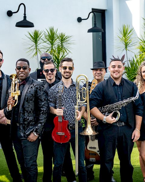 A group of people, each holding musical instruments like a violin, trumpet, ukulele, trombone, saxophone, and a microphone, posing outdoors.