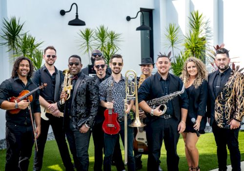 A group of people, each holding musical instruments like a violin, trumpet, ukulele, trombone, saxophone, and a microphone, posing outdoors.