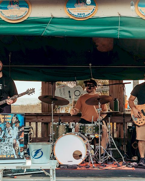 A band is performing on a small outdoor stage, with members playing guitars, drums, and bass, under a green canopy decorated with circular signs.