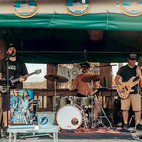 A band is performing on a small outdoor stage, with members playing guitars, drums, and bass, under a green canopy decorated with circular signs.