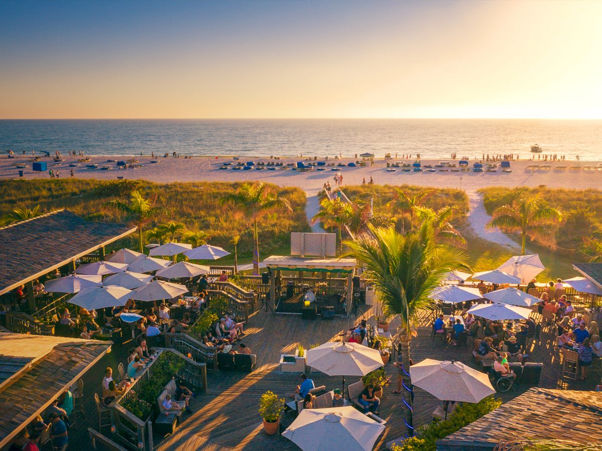 The image shows a vibrant beach scene at sunset, with people seated under umbrellas, palm trees, and the ocean in the background, creating a warm ambiance.
