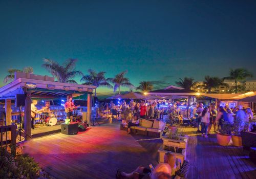A lively outdoor beach bar scene with a band playing on stage, people enjoying the atmosphere, palm trees, and colorful lights at dusk.