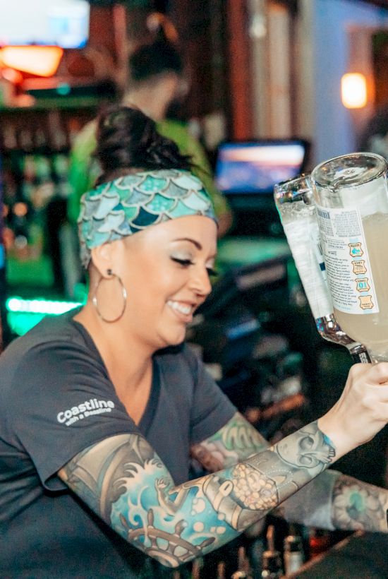 A bartender with tattoos is pouring a drink at a bar surrounded by bottles and bar supplies, while patrons are in the background.