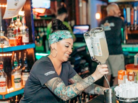 A bartender with tattoos is pouring a drink at a bar, surrounded by bottles and straws on the counter.