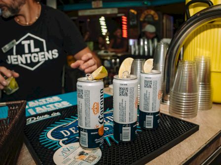 Cans with lime wedges on a bar mat, plastic cups stacked nearby, and a person in a black shirt in the background.