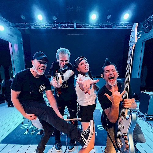 Four people pose energetically on stage, one holding a guitar. They're wearing casual clothes and making playful gestures.