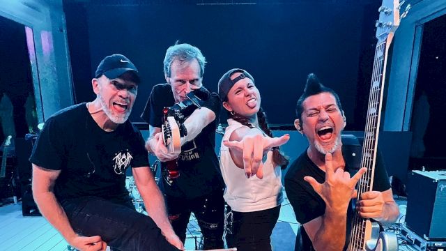 Four people pose energetically on stage, one holding a guitar. They're wearing casual clothes and making playful gestures.