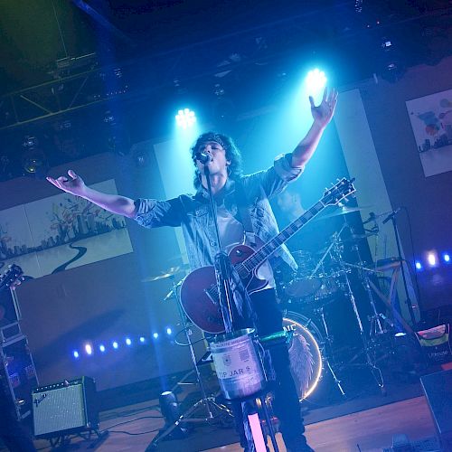 A band is performing on stage with blue lighting; one member has their arms raised, playing guitar, while others play bass and drums.